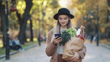 Paket ürünlerin tutarak ve smartphone kullanarak Park'ta yürüyordunuz şık ceket giyen mutlu genç güzel kadın. Alışveriş, sağlıklı beslenme, internet salonu konsept