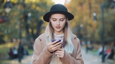 Fashion young woman using smartphone standing in the autumn park. She dressed in coat and hat. Steadicam shot, slow motion