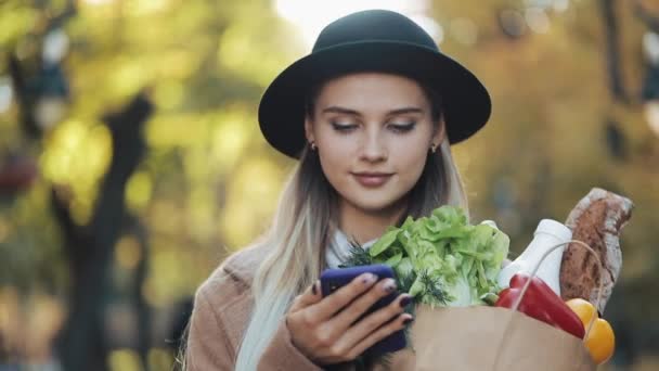행복 한 젊은 아름 다운 여자 들고 제품의 패키지와 스마트폰을 사용 하 여 공원에서 산책 하는 세련 된 코트를 입고. 쇼핑, 건강 한 식습관, 인터넷 쇼핑 개념 — 비디오