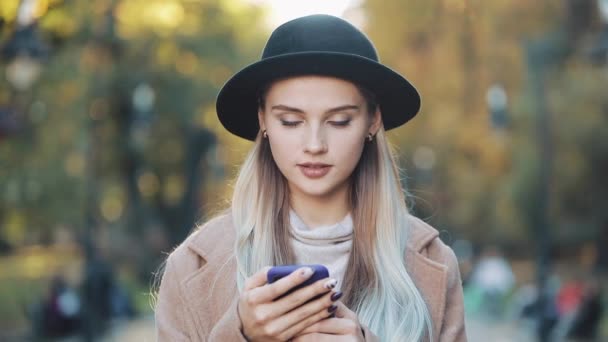 Mode jeune femme utilisant smartphone debout dans le parc d'automne et regardant dans la caméra. Elle portait un manteau et un chapeau. Steadicam shot, ralenti — Video