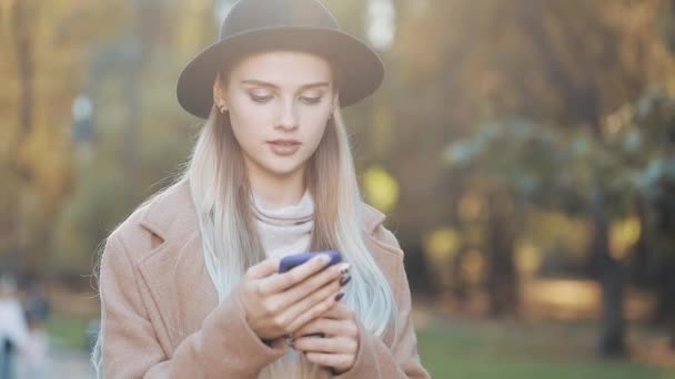 Модная молодая женщина с помощью смартфона стоит в осеннем парке. Она оделась в пальто и шляпу. Стедикам, замедленная съемка — стоковое видео