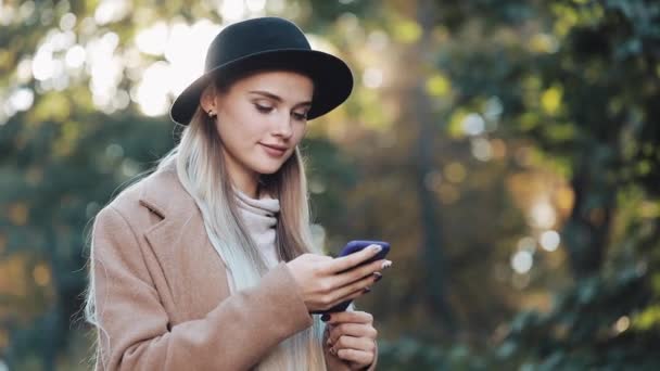 Mode jonge vrouw met behulp van smartphone staande in de herfst park. Ze gekleed in een jas en hoed. Steadicam geschoten, slow-motion. Zonlicht — Stockvideo