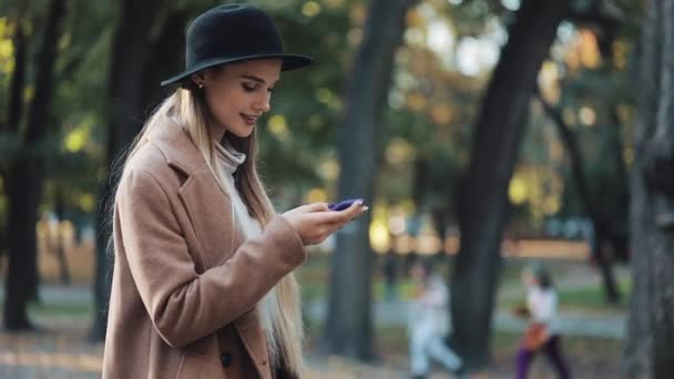 Chica increíble en un aspecto romántico, con sombrero negro caminando por el callejón del parque, mirando a su alrededor y da una sonrisa increíble. Parques, árboles, hojas caídas en el fondo — Vídeos de Stock