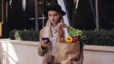 Young beautiful woman wearing stylish coat standing in the street holding package of products and smartphone. She looking into the camera. Shopping, healthy eating, internet-shop concept
