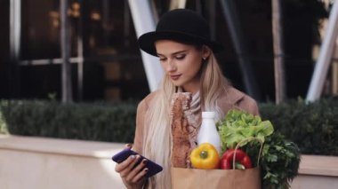Paket ürünlerin tutarak ve smartphone kullanarak sokakta yürürken şık ceket giyen genç güzel kadın. Alışveriş, sağlıklı beslenme, internet salonu konsept