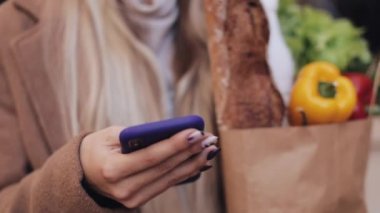 Şık ceket ayakta paket ürünlerin tutarak ve smartphone kullanarak sokakta giyen genç güzel kadın Close-up. Alışveriş, sağlıklı beslenme, internet salonu konsept