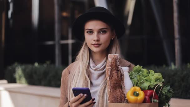 Mladá krásná žena nosí stylový kabát stojící na ulici, drží balíček produktů a smartphone. Kouká do kamery. Nakupování, zdravé stravování, internet-shop koncept — Stock video
