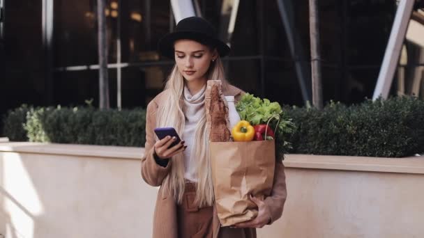 Young beautiful woman wearing stylish coat standing in the street holding package of products and smartphone. She looking into the camera. Shopping, healthy eating, internet-shop concept — 图库视频影像