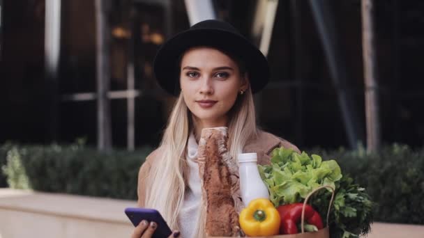 Young beautiful woman wearing stylish coat standing in the street holding package of products and smartphone. She looking into the camera. Shopping, healthy eating, internet-shop concept — 图库视频影像