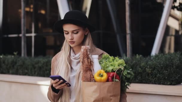 Young beautiful woman wearing stylish coat standing in the street holding package of products and using smartphone. Shopping, healthy eating, internet-shop concept — Stock Video