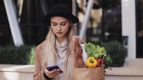 Joven hermosa mujer con un abrigo elegante caminando en la calle sosteniendo el paquete de productos y utilizando el teléfono inteligente. Compras, alimentación saludable, concepto de tienda en Internet — Vídeo de stock