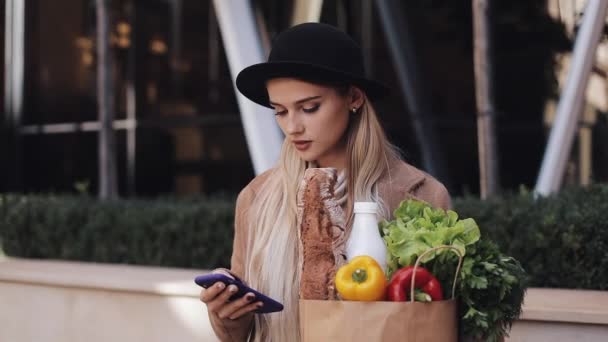Young beautiful woman wearing stylish coat walking in the street holding package of products and using smartphone. Shopping, healthy eating, internet-shop concept — Stock Video