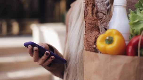 Primer plano de la joven hermosa mujer con un abrigo elegante de pie en la calle que sostiene el paquete de productos y el uso de teléfonos inteligentes. Compras, alimentación saludable, concepto de tienda en Internet — Vídeo de stock