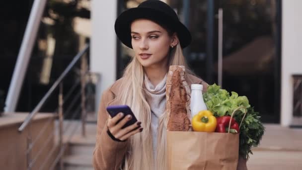 Junge schöne Frau in stylischem Mantel, die auf der Straße mit Produktpaketen herumläuft und das Smartphone benutzt. Einkaufen, gesunde Ernährung, Internet-Shop-Konzept — Stockvideo