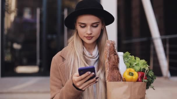 Joven hermosa mujer con un abrigo elegante caminando en la calle sosteniendo el paquete de productos y utilizando el teléfono inteligente. Compras, alimentación saludable, concepto de tienda en Internet — Vídeos de Stock