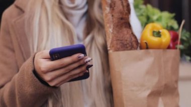 Şık ceket ayakta paket ürünlerin tutarak ve smartphone kullanarak sokakta giyen genç güzel kadın Close-up. Alışveriş, sağlıklı beslenme, internet salonu konsept