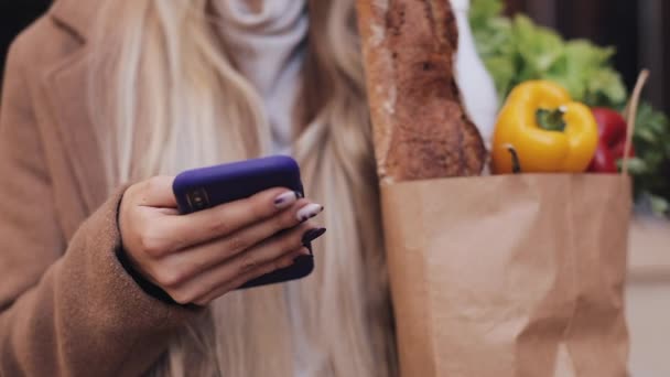 Close-up of young beautiful woman wearing stylish coat standing in the street holding package of products and using smartphone. Shopping, healthy eating, internet-shop concept — Stock Video