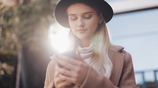 Νεαρή γυναίκα σε παλτό με καπέλο στέκεται κοντά στο Επιχειρηματικό κέντρο της πόλης, επικοινωνεί μέσω smartphone tablet. Steadicam shot, αργή κίνηση — Αρχείο Βίντεο