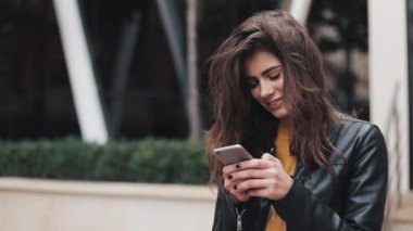 Genç ve güzel kadın kadın deri ceketli telefonunu dururken şehir merkezinde, etrafında görünüyor kullanır. Kentsel binalar arka plan