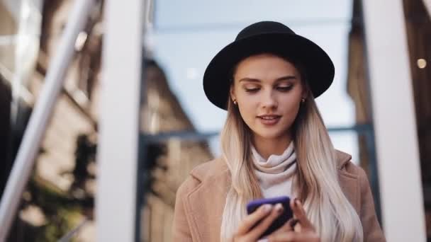 Szczęśliwa Kobieta w płaszcz, kapelusz, chodzenie w pobliżu centrum biznesowego w mieście, komunikuje się za pośrednictwem smartfona, tabletu. Steadicam strzał, zwolnionym tempie — Wideo stockowe