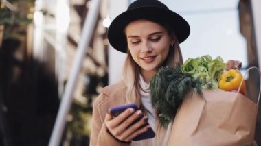 Paket ürünlerin tutarak ve smartphone kullanarak sokakta yürürken şık ceket giyen genç güzel kadın. Alışveriş, sağlıklı beslenme, internet salonu konsept