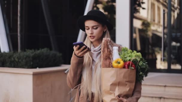 Primer plano de la joven hermosa mujer con un abrigo elegante de pie en la calle sosteniendo el paquete de productos y hablando con el teléfono inteligente. Ella escucha el mensaje de voz — Vídeo de stock