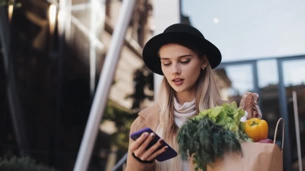 Joven hermosa mujer con un abrigo elegante caminando en la calle sosteniendo el paquete de productos y utilizando el teléfono inteligente. Compras, alimentación saludable, concepto de tienda en Internet — Vídeos de Stock