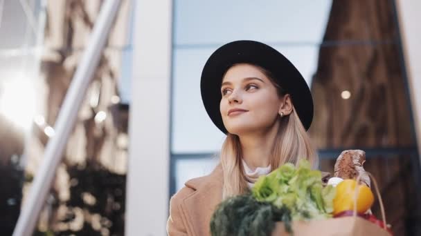 Mujer hermosa joven con un abrigo elegante caminando por la calle sosteniendo un paquete de productos sonriendo y mirando a su alrededor. Compras, alimentación saludable — Vídeos de Stock