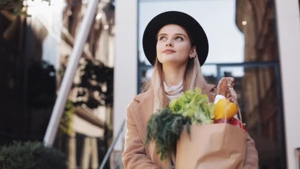 Mujer hermosa joven con un abrigo elegante caminando por la calle sosteniendo un paquete de productos sonriendo y mirando a su alrededor. Compras, alimentación saludable — Vídeos de Stock