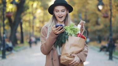 Paket ürünlerin tutarak ve smartphone kullanarak sokakta yürürken şık ceket giyen genç güzel kadın. Alışveriş, sağlıklı beslenme, internet salonu konsept