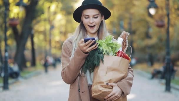 Jeune belle femme portant un manteau élégant marchant dans la rue tenant paquet de produits et en utilisant un smartphone. Shopping, alimentation saine, concept internet-shop — Video