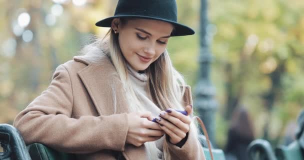 Mulher bonita de casaco usando smartphone relaxa no banco no parque de outono. Tecnologia ao ar livre — Vídeo de Stock