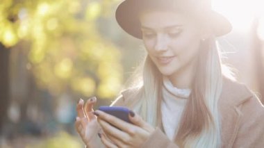 Smartphone ayakta sonbahar parkta kullanarak bir ceket giyen genç kadın. Teknoloji açık havada. Güneş ışığı.