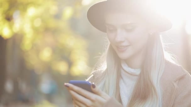 Jovem mulher vestindo um casaco usando smartphone em pé no parque de outono. Tecnologia ao ar livre. Luz solar — Vídeo de Stock