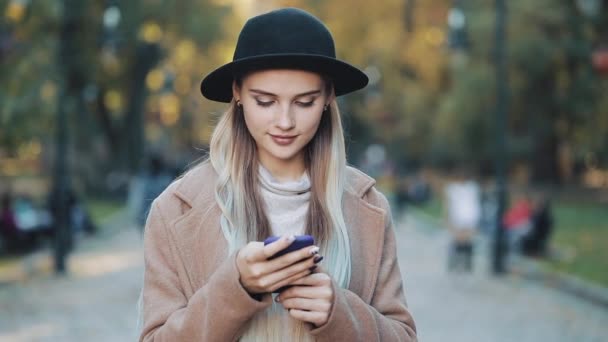 Moda giovane donna utilizzando smartphone in piedi nel parco autunnale e guardando nella fotocamera. Si è vestita con cappotto e cappello. Colpo di Steadicam, rallentatore. Luce del sole — Video Stock