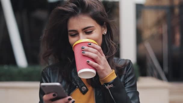 Молода щаслива жінка використовує смартфон і п'є каву, щоб вийти на вулицю, йдучи по міській вулиці, повільний рух — стокове відео