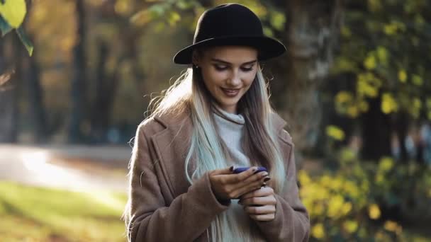 Mujer joven con un abrigo usando un teléfono inteligente de pie en el parque de otoño. Tecnología al aire libre. Luz solar — Vídeo de stock