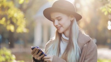 Smartphone ayakta sonbahar parkta kullanarak bir ceket giyen genç kadın. Teknoloji açık havada. Güneş ışığı