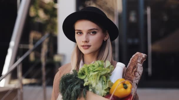 Jeune belle femme portant un manteau élégant debout dans la rue urbaine tenant un paquet de produits souriant et regardant dans la caméra. Shopping, alimentation saine — Video