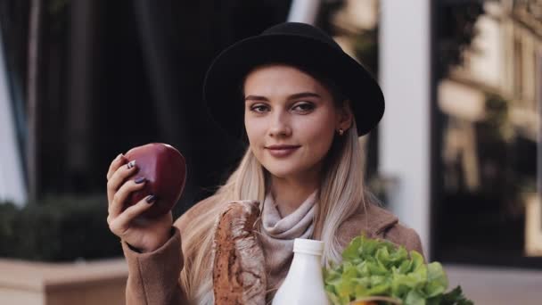 Молодая красивая женщина в стильном пальто стоит на улице с пакетом продуктов, улыбаясь и глядя в камеру. Она держит красное яблоко в руке. Шоппинг, здоровое питание — стоковое видео