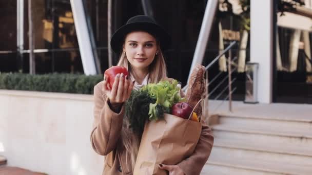 Mujer hermosa joven con un abrigo elegante de pie en la calle sosteniendo un paquete de productos sonriendo y mirando a la cámara. Ella sostiene el pimiento rojo en su mano. Compras, alimentación saludable — Vídeos de Stock