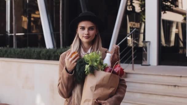 Mujer hermosa joven con un abrigo elegante de pie en la calle sosteniendo un paquete de productos sonriendo y mirando a la cámara. Ella sostiene el pimiento amarillo en su mano. Compras, alimentación saludable — Vídeos de Stock