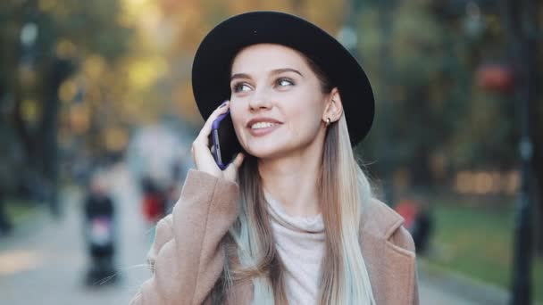 Ung vacker kvinna talar på hennes mobiltelefon medan promenader i parken hösten på en bakgrund av gula blad. Flicka klädd i kappa med hjälp av sin smartphone — Stockvideo