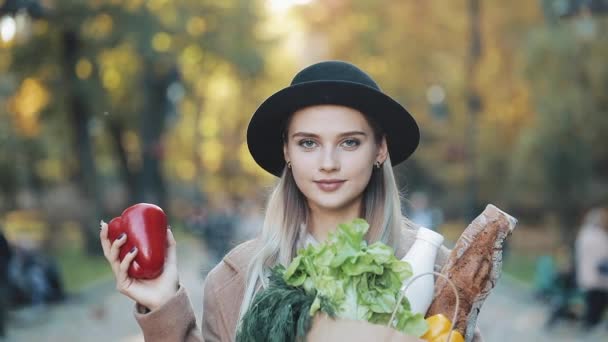 Young beautiful woman wearing stylish coat standing in the park holding a package of products smiling and looking into the camera. She holds the red pepper in her hand. Shopping, healthy eating — Stock Video