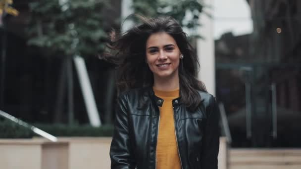 Pretty young woman in the leather jacket is walking near the business center. The girl fixes her hair looking into the camera — Stock Video
