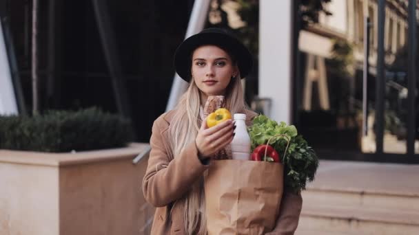 Mujer hermosa joven con un abrigo elegante de pie en la calle sosteniendo un paquete de productos. Ella sostiene el pimiento amarillo en su mano. Compras, alimentación saludable, productos frescos — Vídeos de Stock