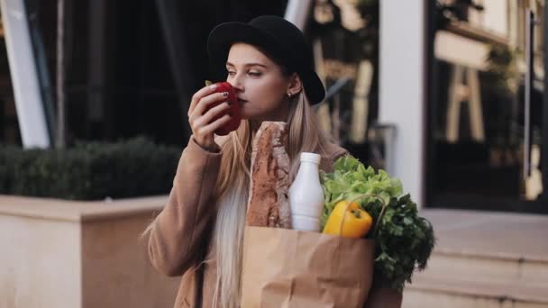 Mujer hermosa joven con un abrigo elegante de pie en la calle sosteniendo un paquete de productos. Ella sostiene la manzana roja en su mano. Compras, alimentación saludable, productos frescos — Vídeos de Stock