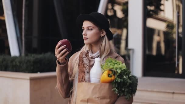 Молодая красивая женщина в стильном пальто стоит на улице с пакетом продуктов. Она держит красное яблоко в руке. Шоппинг, здоровое питание, свежие продукты — стоковое видео