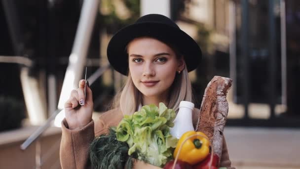 Joven hermosa mujer con un abrigo elegante de pie en la calle con un paquete de productos y tiene tarjeta de crédito. Compras, alimentación saludable, concepto de tienda en Internet — Vídeo de stock