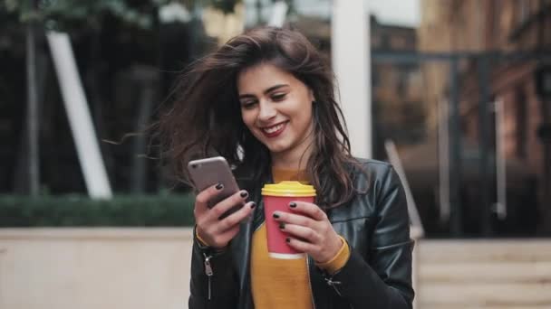 Wanita muda yang bahagia menggunakan smartphone dan minum kopi untuk pergi keluar ruangan, berjalan menyusuri jalan kota, gerakan lambat — Stok Video